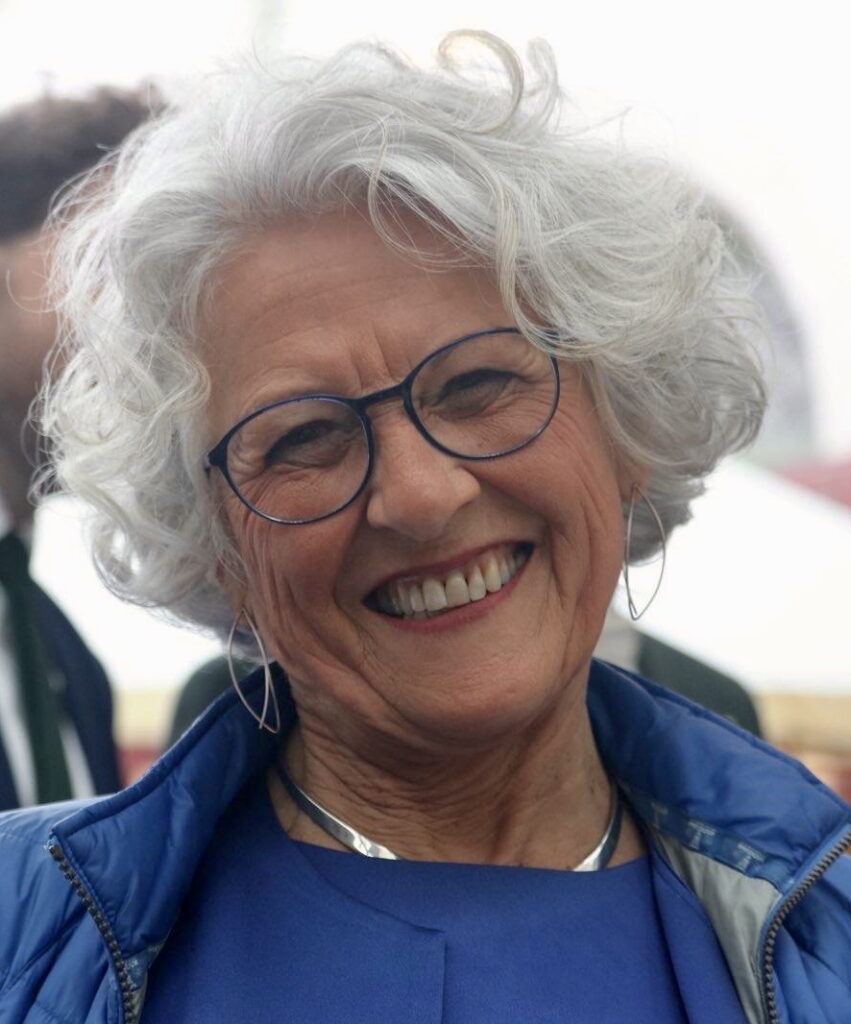 Lächelnde ältere Frau mit kurzen, lockigen grauen Haaren und Brille, die eine blaue Jacke und silberne Ohrringe trägt, steht im Freien.