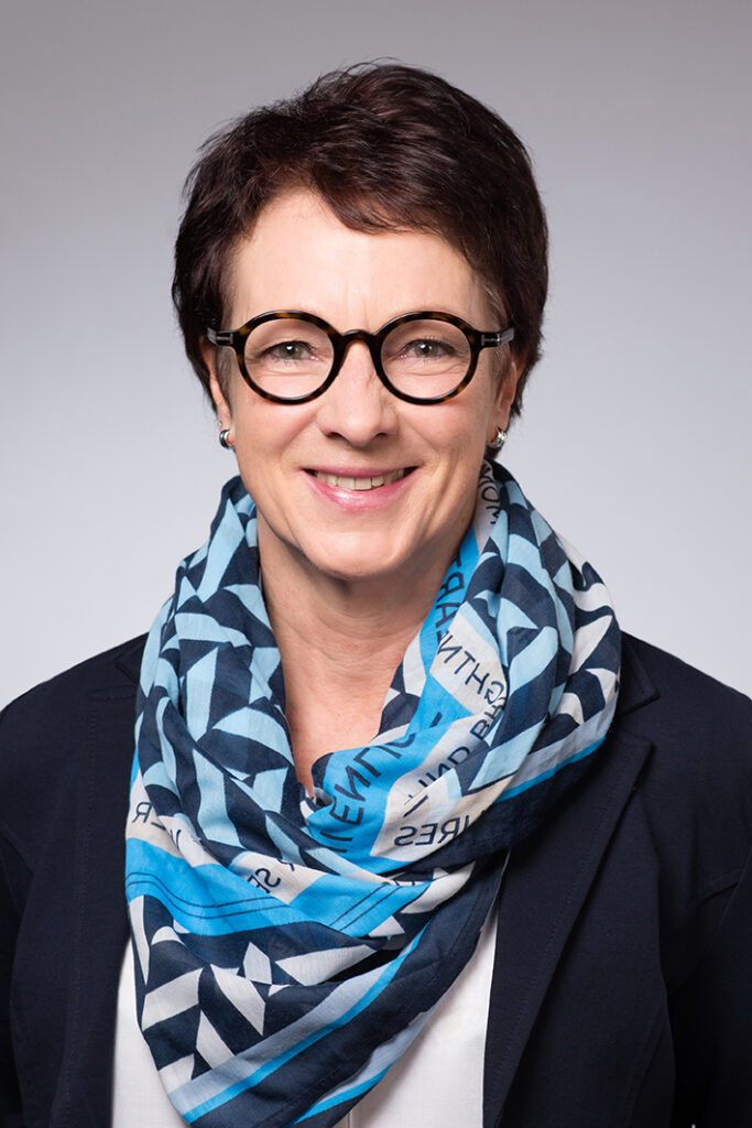 Eine Frau mit kurzen dunklen Haaren und einer runden Brille lächelt in die Kamera. Sie trägt einen dunklen Blazer und einen blau gemusterten Schal mit Text vor einem schlichten hellgrauen Hintergrund.