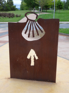 Ein Metallschild mit einem Muschelausschnitt und einem aufgemalten Pfeil, der nach vorne zeigt und eine Richtung auf dem Jakobsweg angibt. Im Hintergrund sind grünes Gras, Bäume und eine Straße zu sehen.