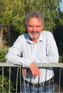 Ein lächelnder älterer Mann mit grauem Haar und Bart lehnt an einem Metallzaun in einem sonnigen Garten, er trägt ein hellblaues Button-up-Hemd und Jeans. Im Hintergrund sind grüne Bäume und Pflanzen zu sehen.