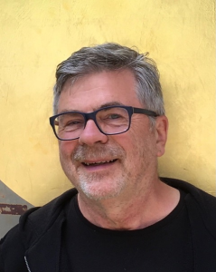 Ein Mann mittleren Alters mit kurzen grauen Haaren, Brille und Bart lächelt. Er trägt ein schwarzes Hemd und eine schwarze Jacke und steht vor einem goldfarbenen Hintergrund.