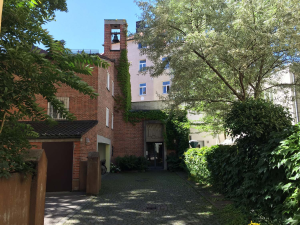 Sonniger Innenhof mit einem von Efeu überwucherten Backsteingebäude, einem Garagentor auf der linken Seite, einem weißen Gebäude im Hintergrund und üppigen grünen Bäumen und Sträuchern in der Umgebung.