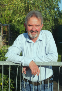 Ein älterer Mann mit grauem Haar und Bart lächelt, während er sich im Freien an einen Metallzaun lehnt, mit grünen Bäumen und Sonnenlicht im Hintergrund. Er trägt ein hellblaues, langärmeliges Button-up-Hemd und Jeans.
