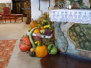 Eine farbenfrohe Herbstdekoration mit Kürbissen, Kürbissen und Blumen ist neben einem mit weißem Spitzentuch drapierten Kirchenaltar aufgebaut. Die Kulisse besteht aus verzierten Stühlen und einem gemusterten Teppich im Hintergrund.