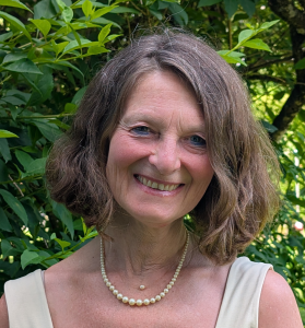 Eine Frau mit schulterlangen braunen Haaren, die ein cremefarbenes ärmelloses Oberteil und eine Perlenkette trägt, steht lächelnd vor einem grünen Laubgebüsch.