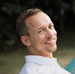 Ein Mann mit kurzen hellbraunen Haaren und einem gestutzten Spitzbart lächelt und trägt ein weißes Hemd mit Kragen. Er befindet sich im Freien, mit einem unscharfen Hintergrund aus Grünpflanzen und Bäumen.