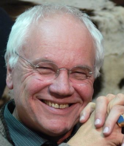 Ein älterer Mann mit kurzen weißen Haaren und Brille lächelt warm, die Augen geschlossen, die Hände vor dem Gesicht verschränkt. Er trägt ein dunkles Hemd und eine gemusterte Jacke.
