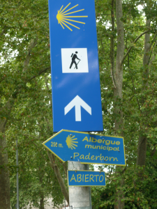 Blaue Schilder mit gelben Muschelsymbolen markieren den Jakobsweg; eines weist nach vorne, ein anderes nach links zu einer 250 Meter entfernten offenen städtischen Herberge in Paderborn. Grüne Bäume füllen den Hintergrund.