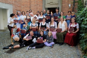 Eine große Gruppe von Menschen, viele in traditioneller bayerischer Kleidung, posiert im Freien auf Kopfsteinpflaster vor einem Backsteingebäude, hält braune Mappen in der Hand und lächelt in die Kamera. Einige Kinder sitzen ganz vorne.