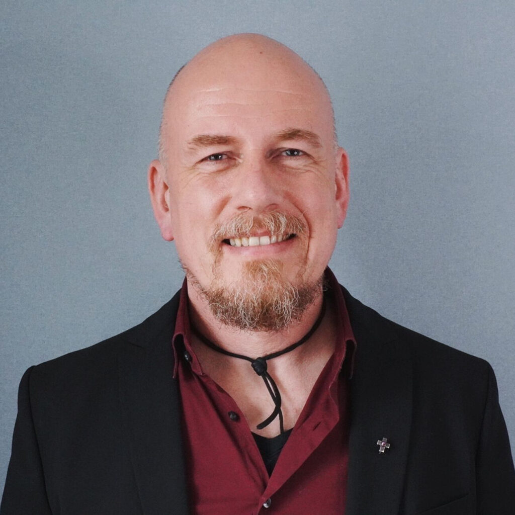Ein lächelnder Mann mit Glatze und Spitzbart, der einen schwarzen Blazer über einem weinroten Hemd trägt, mit einer Bolo-Krawatte und einer Anstecknadel am Revers, steht vor einem schlichten grauen Hintergrund.