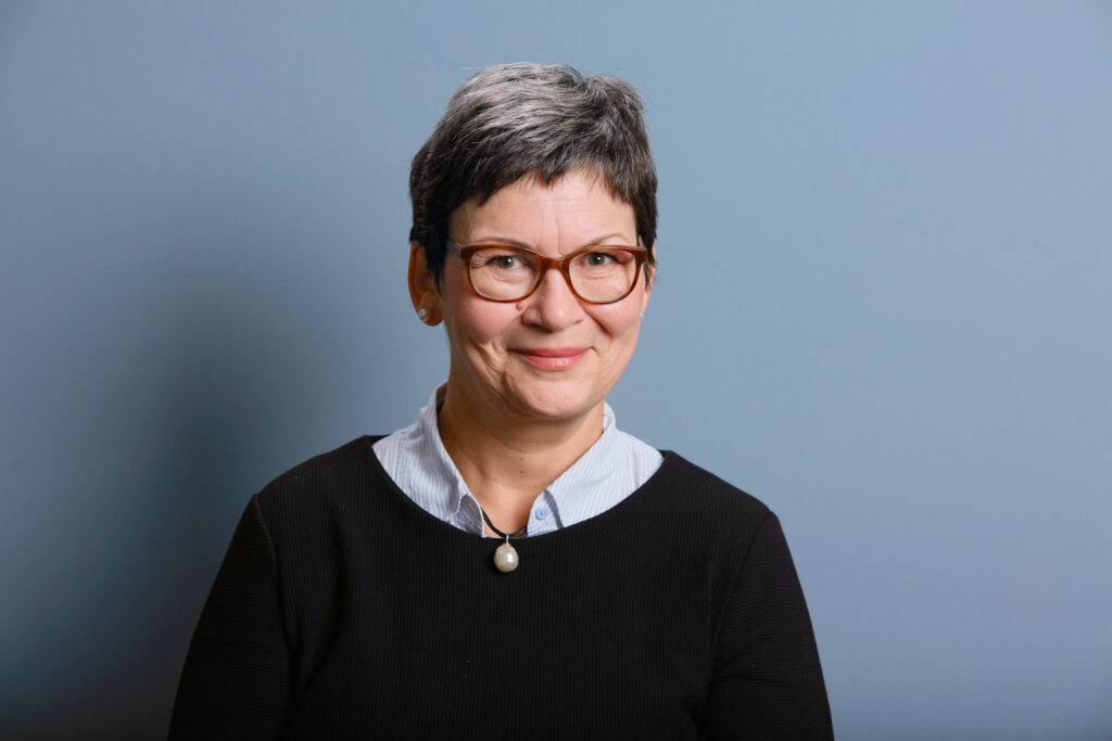 Eine Frau mit kurzen grauen Haaren und Brille lächelt. Sie trägt einen schwarzen Pullover über einem Hemd mit Kragen und eine Perlenkette und steht vor einem einfarbig blauen Hintergrund.