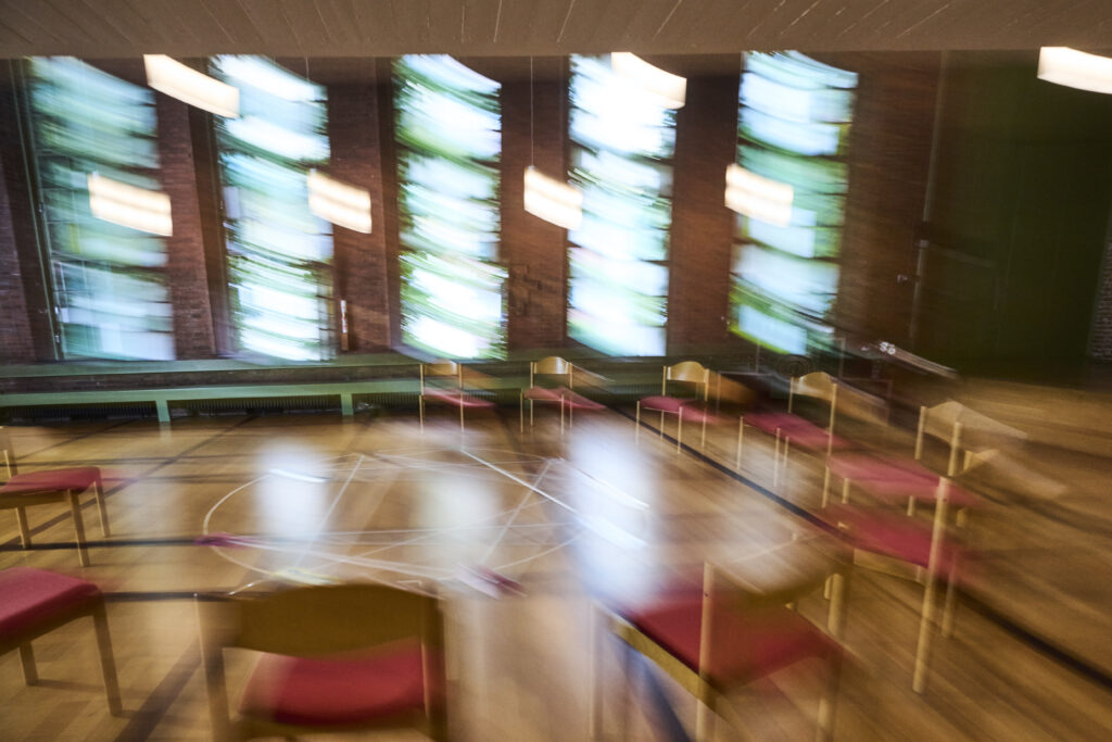 Unscharfes Bild eines leeren Raums mit kreisförmig angeordneten Holzstühlen auf einem Holzboden; hohe Fenster im Hintergrund lassen Licht herein, das durch die Bewegungsunschärfe Schlieren erzeugt.