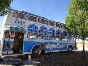 Ein zum Café umgebauter Doppeldeckerbus ist unter Bäumen geparkt. Der Bus hat blau-weiß gestrichene Markisen an den Fenstern und die Aufschrift Café und Doppellecker an der Seite. Eine Person mit einem Rucksack steht in der Nähe.