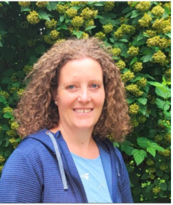 Eine Frau mit lockigem braunem Haar, die einen blauen Kapuzenpullover mit Reißverschluss und ein hellblaues Hemd trägt, steht lächelnd vor einem belaubten Busch mit kleinen gelbgrünen Blütenbüscheln.