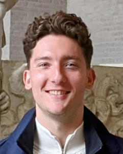 Ein junger Mann mit kurzen, lockigen braunen Haaren lächelt in die Kamera. Er trägt eine dunkle Jacke über einem hellen Hemd und steht in einem Innenraum mit einer Steinmauer und Skulpturen im Hintergrund.