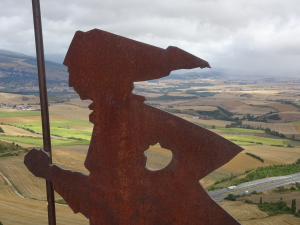 Die Metallsilhouette eines Pilgers, der einen Stab in der Hand hält, überblickt eine weite ländliche Landschaft mit Feldern und fernen Hügeln unter einem bewölkten Himmel.