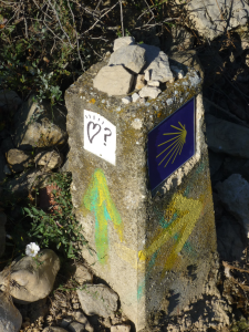 Betonstein mit einem Muschelsymbol, einem weißen Aufkleber mit einem Herz und einem Fragezeichen, grün und gelb gemalten Pfeilen, Steinen auf der Spitze, umgeben von Gras und Steinen.