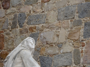 Eine steinerne Statue einer verhüllten Figur steht vor einer strukturierten Steinmauer aus verschieden geformten und farbigen Steinen.
