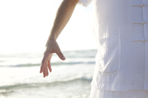 Eine weiß gekleidete Person steht mit einem nach unten ausgestreckten Arm am Meer, während sich das Sonnenlicht auf dem Wasser im Hintergrund spiegelt.