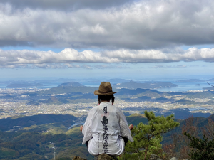 Eine Person, die einen Strohhut und ein weißes Gewand trägt, sitzt auf einem Berggipfel und überblickt eine weite Landschaft mit Hügeln, einer Stadt und dem Meer unter einem bewölkten Himmel.