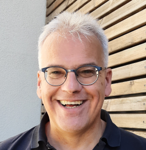Ein lächelnder älterer Mann mit kurzen weißen Haaren und Brille, der ein dunkles Poloshirt trägt, steht vor einer Holzlattenwand und einer hellen Wand.