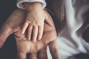 Eine kleine Babyhand, die auf der offenen Handfläche eines Erwachsenen ruht, zeigt den Größenunterschied und symbolisiert Fürsorge, Schutz und Verbindung. Beide Hände sind weich fokussiert und sanft beleuchtet.