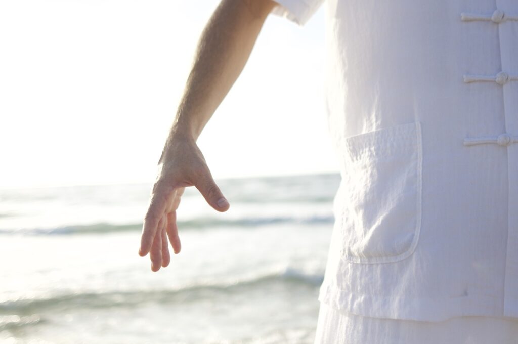 Eine weiß gekleidete Person steht am Meer, einen Arm nach unten gestreckt, die Finger entspannt. Der Hintergrund ist hell und unscharf und zeigt sanfte Wellen und einen sonnenbeschienenen Himmel.
