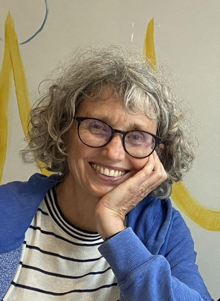 Eine ältere Frau mit lockigem, grauem Haar und Brille lächelt und stützt ihr Kinn auf ihre Hand. Sie trägt ein gestreiftes Hemd und eine blaue Jacke, im Hintergrund ist eine bemalte Wand zu sehen.