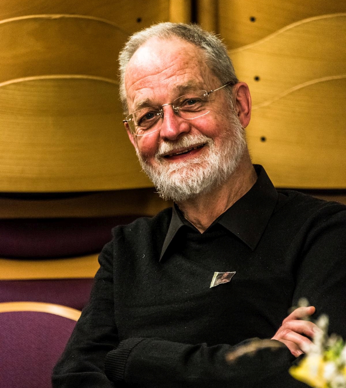 Ein älterer Mann mit Brille, weißem Bart und schwarzem Hemd lächelt, während er mit verschränkten Armen vor hölzernen und lila Stühlen sitzt.