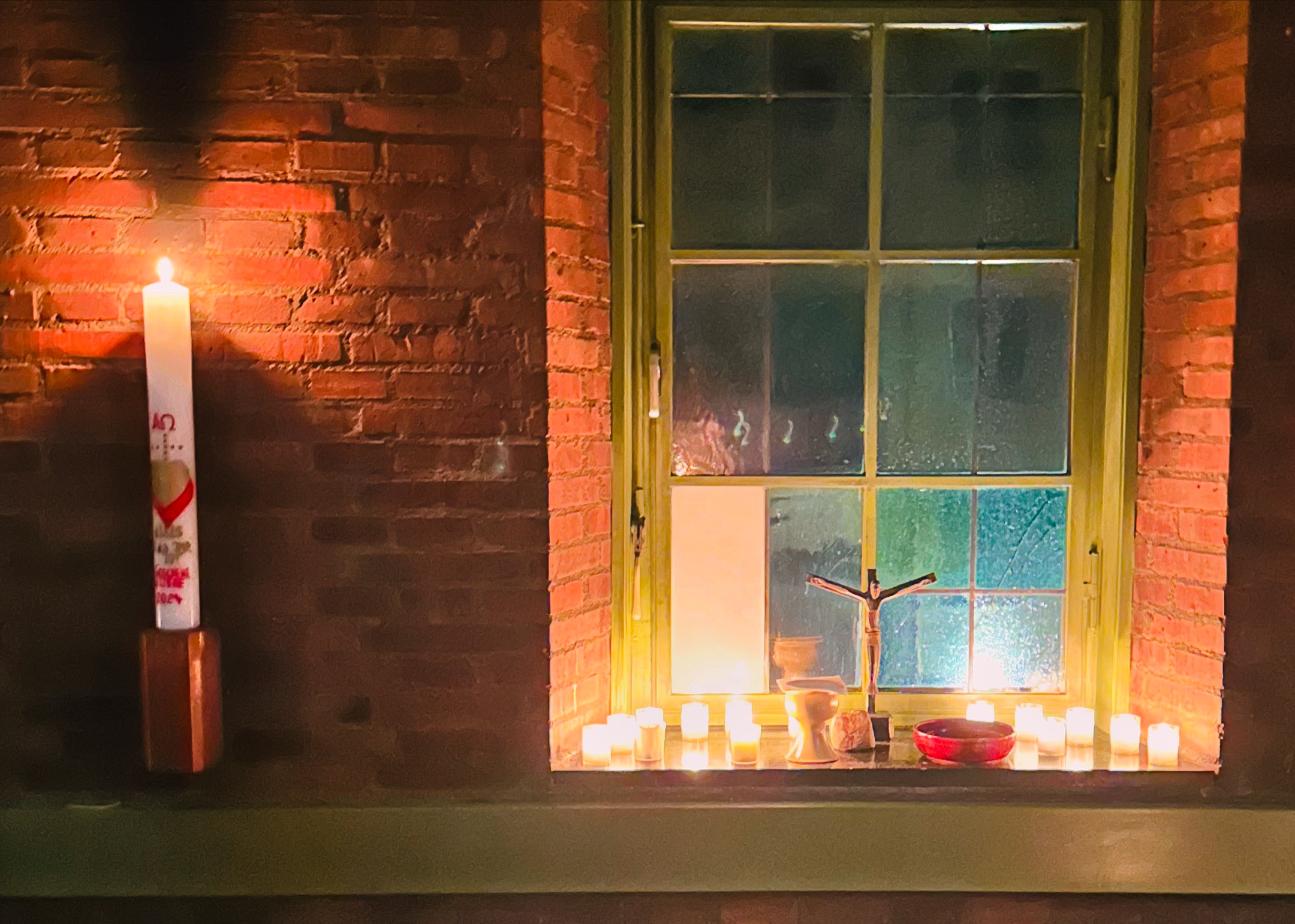 Eine brennende Kerze an einer Backsteinwand und mehrere kleine Kerzen auf einem Fenstersims neben einem Kruzifix, einem Kelch und einer Schale schaffen eine warme, friedliche Atmosphäre.