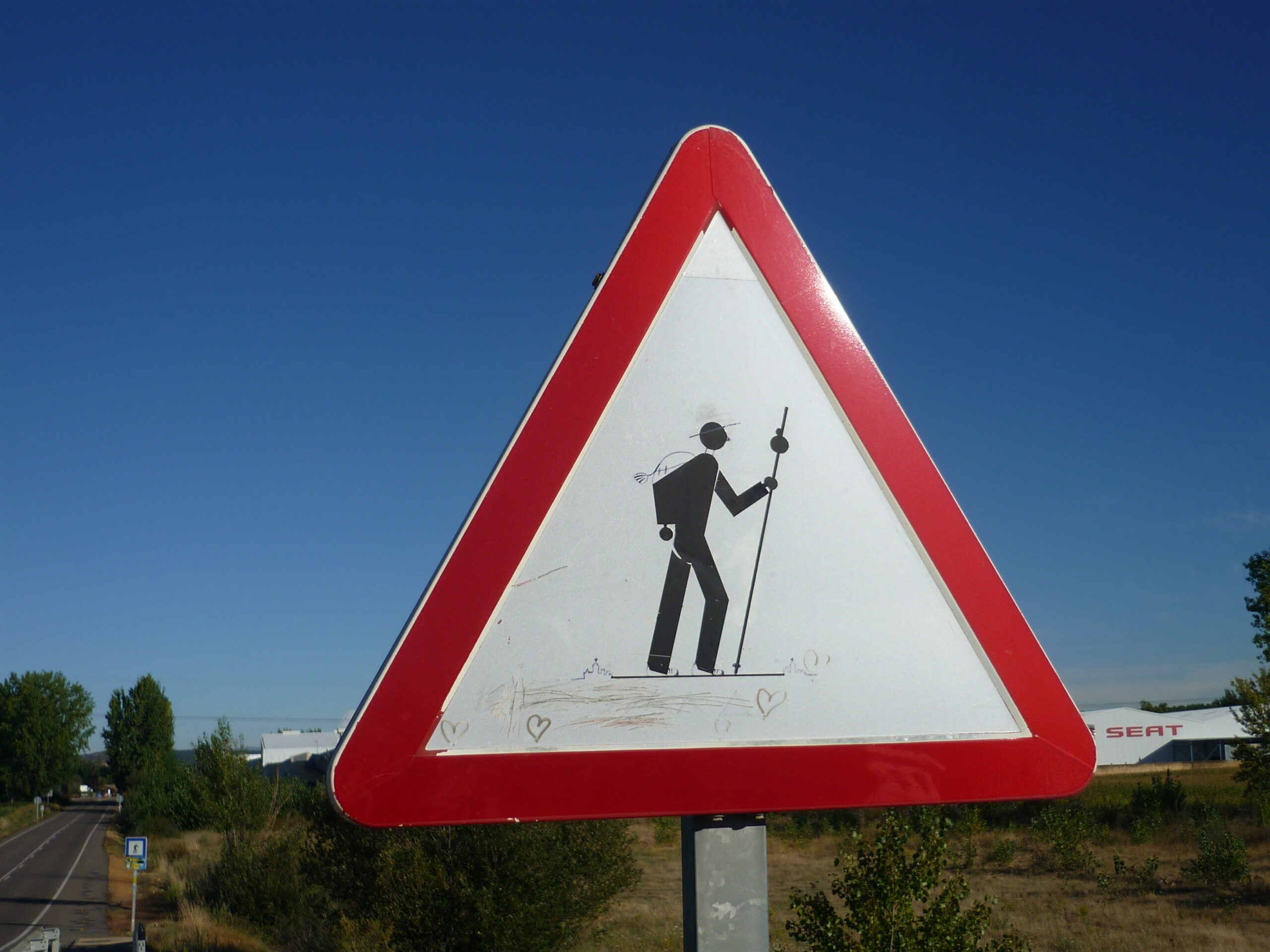 Ein dreieckiges Straßenschild mit rotem Rand zeigt ein Strichmännchen mit Rucksack und Wanderstock, das über das Originalschild gezeichnet wurde. Klarer blauer Himmel im Hintergrund.