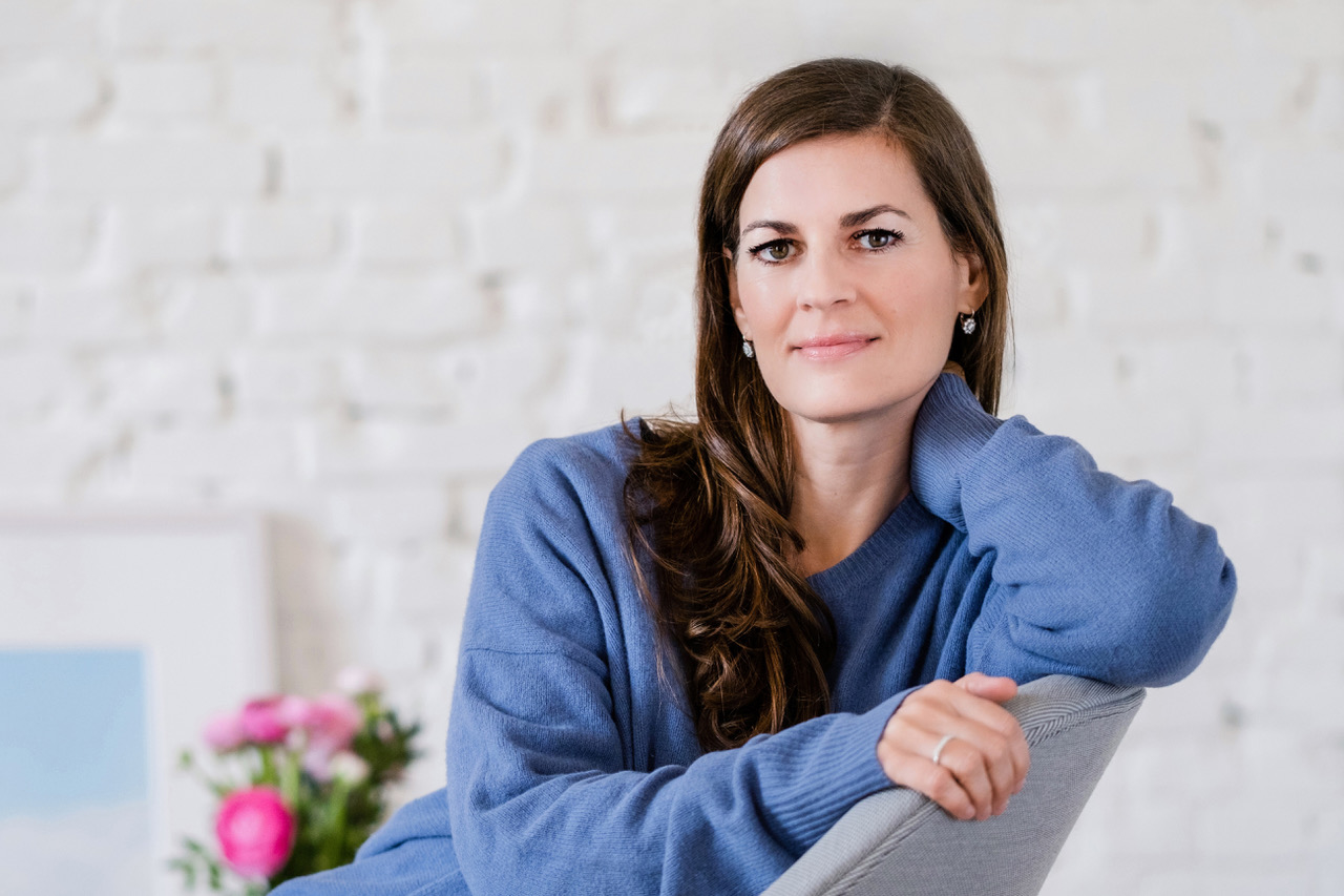 Frau mit langen braunen Haaren und blauem Pullover, auf einem grauen Stuhl sitzend, sanft lächelnd, eine Hand auf den Kopf gestützt; weiße Backsteinmauer und Blumen im Hintergrund.