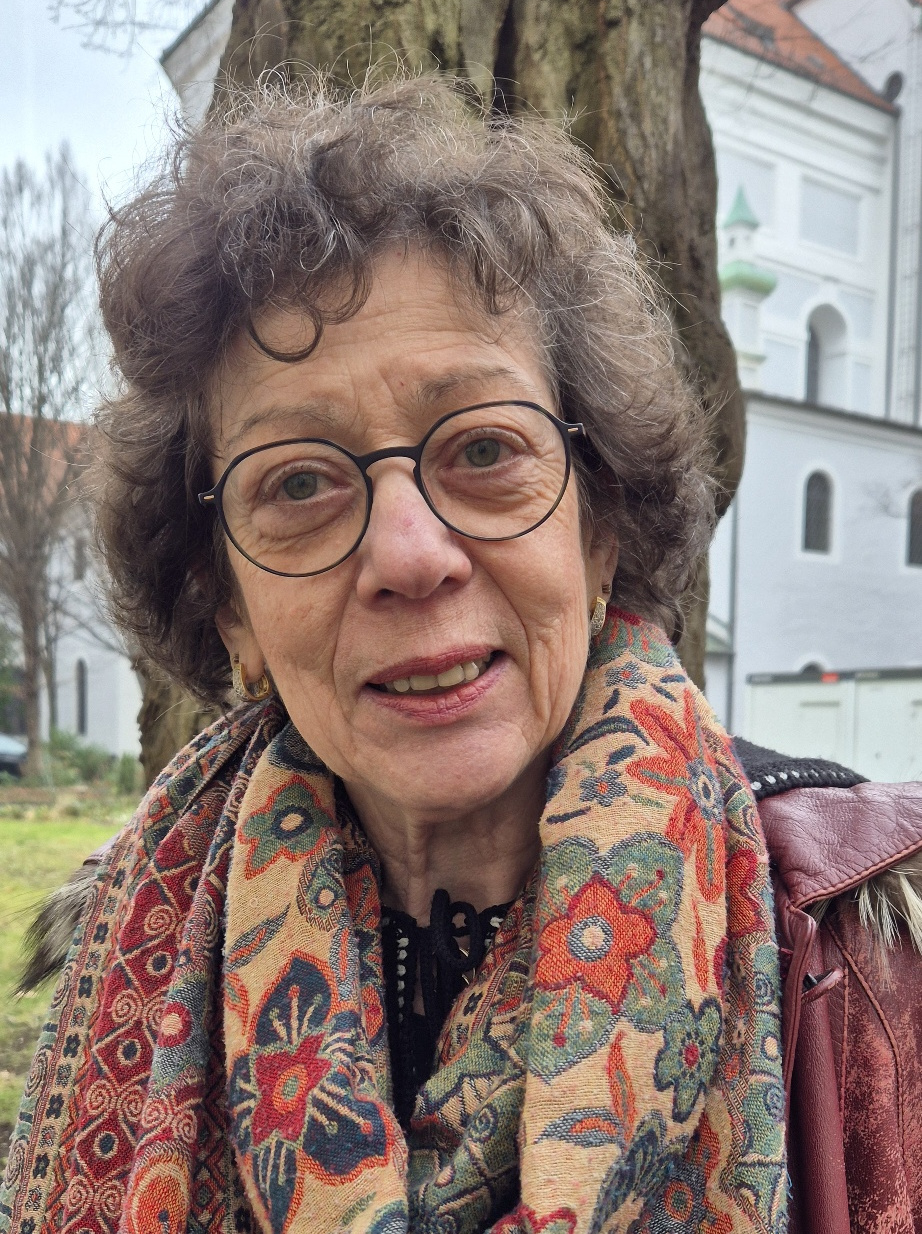 Eine ältere Frau mit lockigem Haar und Brille steht im Freien an einem Baum. Sie trägt einen bunt gemusterten Schal und eine kastanienbraune Jacke, im Hintergrund ist ein Gebäude zu sehen.