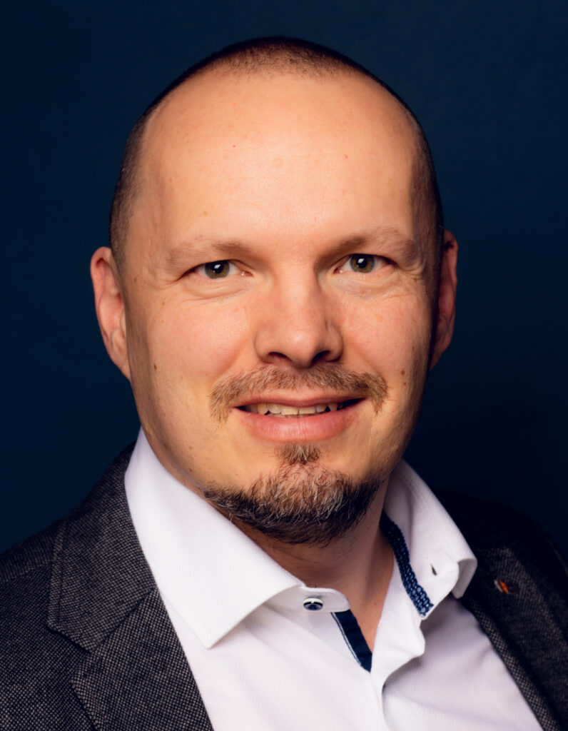 Ein Mann mit gestutztem Bart und Schnurrbart, der ein weißes Hemd und einen grauen Blazer trägt, lächelt in die Kamera vor einem dunkelblauen Hintergrund.