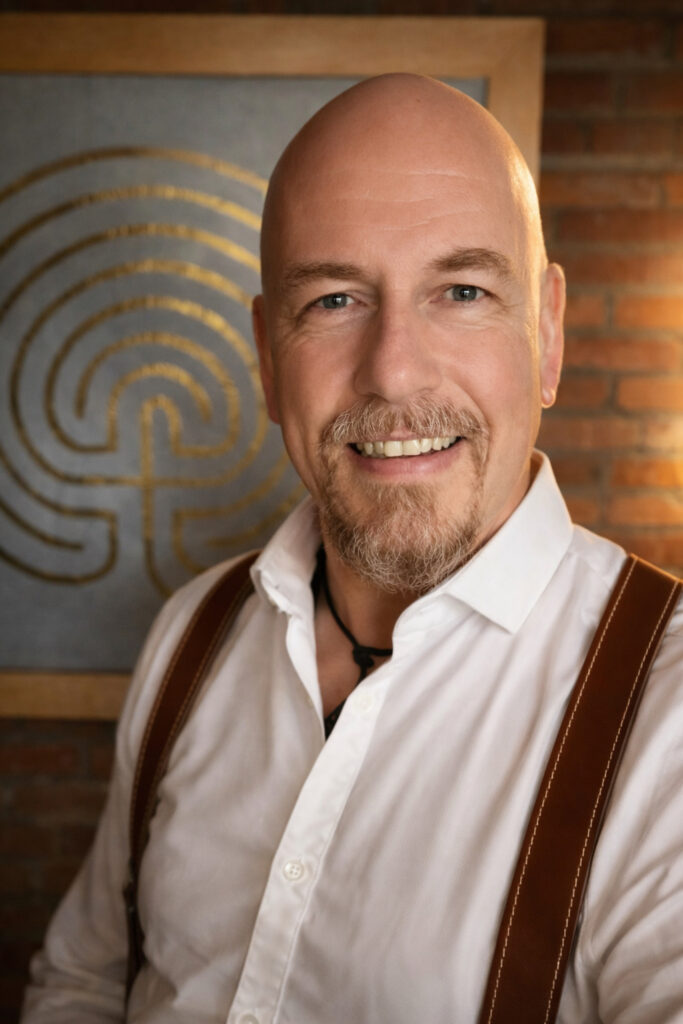 Ein lächelnder Mann mit Glatze und Spitzbart, der ein weißes Hemd und braune Hosenträger trägt, steht vor einer Backsteinmauer und abstrakter Kunst mit einem goldenen Labyrinthmuster.